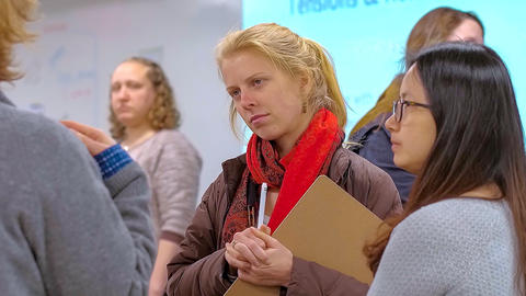 Close up of graduate student in a small group of other students, with clip board and pen, head tilted and listening.