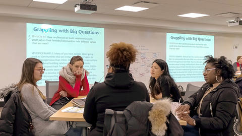 Five graduate students seated at a table in discussion. Projected on the screen behind them are slides that read "Grappling with Big Questions" and then smaller texts beneath.