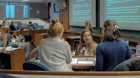 Close up of three female graduate students engaged in discussion as small groups work behind them in the classroom.