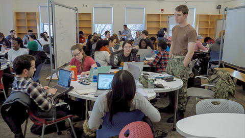 Classroom of students working at small round tables on computers.