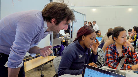 Medium close up of Teaching Assistant leaning over the computer of a student in a classroom full of students