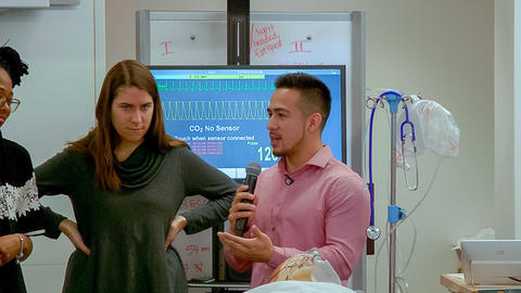Close up of two graduate students, one speaking into a microphone. Around the students are medical monitors and machines.