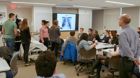 Wide shot of classroom with some students seated and some students gathered around a fake patient on a hospital bed hooked up to machines.