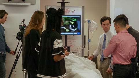 Medium close up of a group of students in a classroom gathered around a pretend patient on a hospital bed, hooked up to monitors.