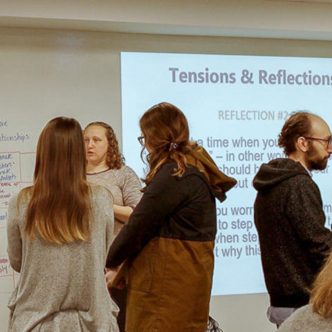 Graudate standing in groups of 4 discussing. Slide projected on screen behind them reads "Tensions & Reflections."