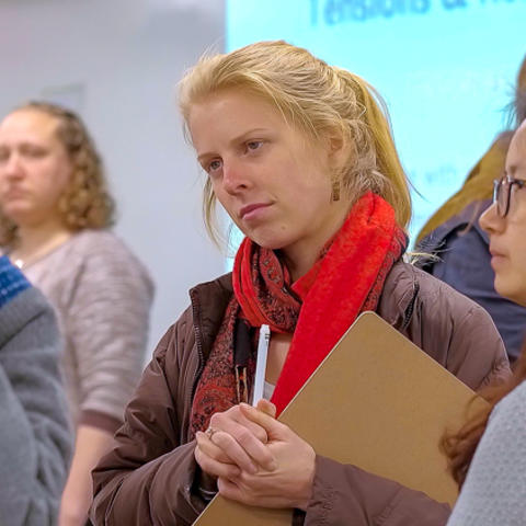 Close up of graduate student in a small group of other students, with clip board and pen, head tilted and listening.