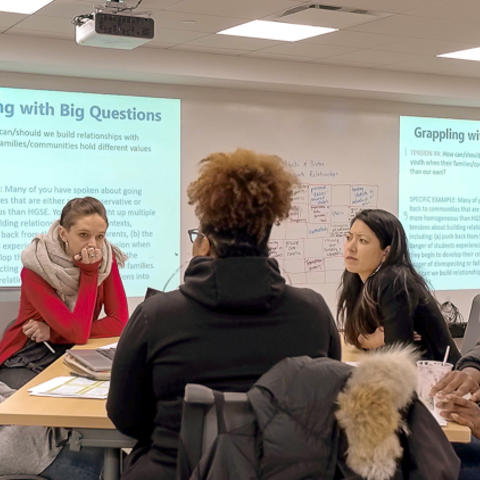 Five graduate students seated at a table in discussion. Projected on the screen behind them are slides that read "Grappling with Big Questions" and then smaller texts beneath.