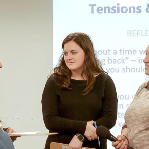 Four graduate students and Instructor Brion-Meisels standing together in a small group in discussion. There is a section of a projected slide visible behind them that reads "Tension & Reflections."