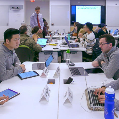 Wide shot of four graduate students seated at a table facing eachother, 2 with tablets, 2 with computers. Around them the rest of the class is seated in similar formation.