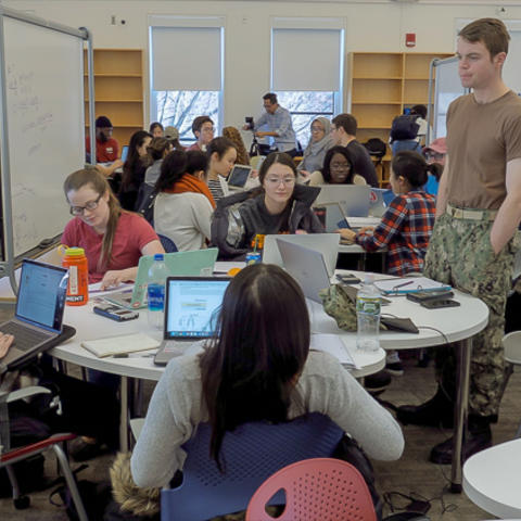 Classroom of students working at small round tables on computers.